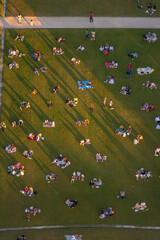 Drone view of groups of people, their long shadows projected onto the green lawns of the Champ de Mars at sunset, relaxing, enjoying picnics and romantic dinners.