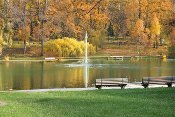 Autumn park scene with a central water fountain in a calm lake, surrounded by colorful trees and benches on a green lawn.