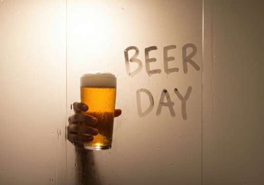 Woman holding a glass of frothy beer behind a steamy glass with "beer day" written on it, celebrating the holiday.