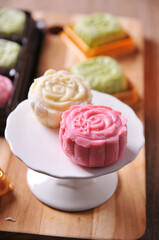 Two delicate snow skin mooncakes, one pink and one white, are elegantly displayed on a small white cake stand, with more mooncakes blurred in the background on a wooden surface