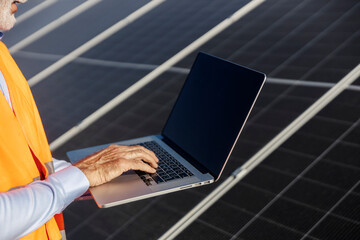 Close up of senior supervisor standing with laptop near solar panels and testing them.