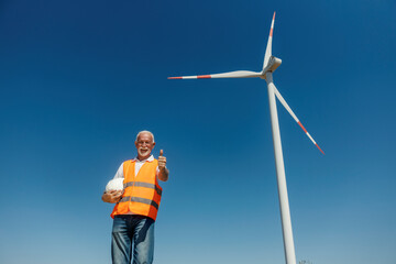 An old engineer standing near wind turbine and giving thumbs up at the camera.