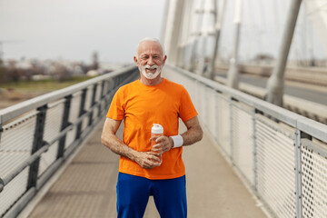 Portrait of senior sportsman in shape standing on a bridge with bottle of water in hands and...