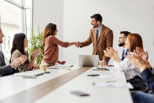 Happy businesswoman shaking hands with ceo while leadership team sitting at conference table and applauding.