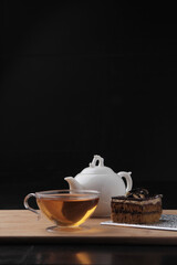A clear teacup filled with amber tea sits next to a white teapot and a slice of chocolate cake on a...