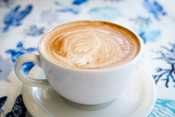 A top-down shot of a delicious cappuccino with detailed latte art, presented in a white cup on a table with a beautiful blue and white marine pattern.