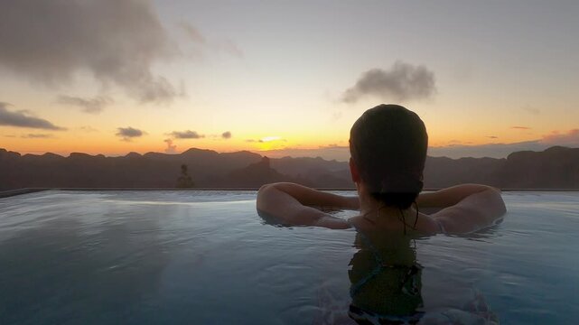Woman in her 30's relaxing in a pool, leaning on the edge while watching a serene natural sunset over distant mountains with space for text on side