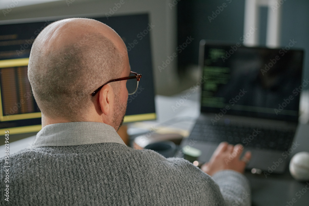 Poster middle aged caucasian man working on laptop with code visible on screen, sitting at desk in modern o