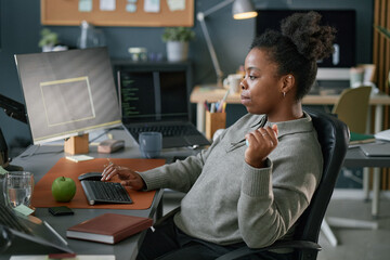 Young adult Black woman working at computer in modern office, sitting at desk using hand exerciser...