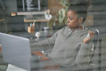 Young adult Black woman working at desk using laptop and holding grip trainer, focusing on computer...