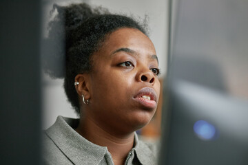 Portrait of young adult Black woman looking at computer screen with focused expression, visible...