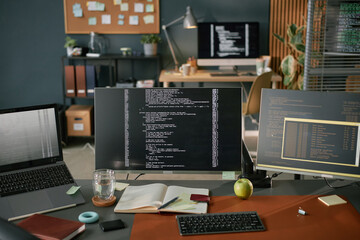 Three computer monitors displaying programming code on desk with open notebook, pen, glass of...