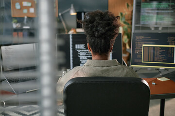 Back view of young Black woman sitting at desk working on computer with multiple monitors...