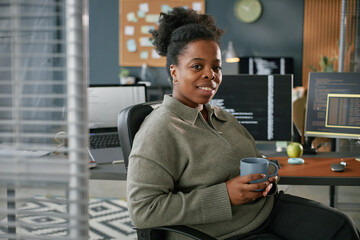Portrait of young adult Black woman sitting at desk in modern office holding mug, smiling at camera...