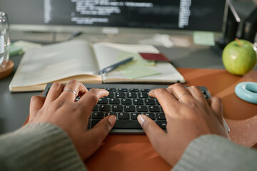 Black woman typing on wireless keyboard with both hands, working at cluttered desk with notebook,...