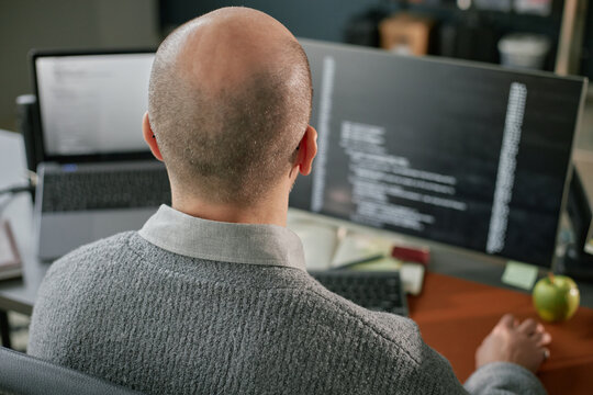 Middle aged Caucasian man sitting at desk working on computer monitors with programming code visible on screens, back of head and shoulders in focus, office environment
