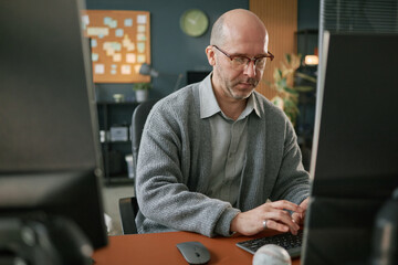 Middle aged Caucasian man wearing glasses working on computer in modern office, sitting at desk and...
