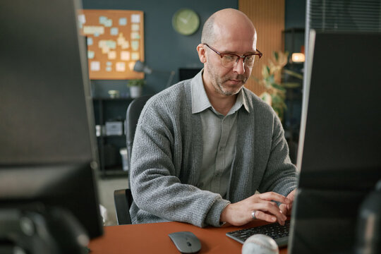 Middle aged Caucasian man wearing glasses working on computer in modern office, sitting at desk and typing on keyboard, focused expression, office background with clock and notes