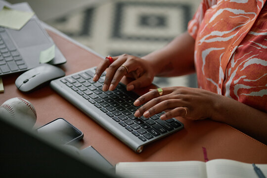 Black woman typing on wireless keyboard at desk, hands visible with rings, working near laptop, smartphone, notebook and baseball, suggesting multitasking in modern workspace - Powered by Adobe