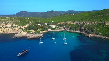 sailboats and yachts moored in turquoise waters of a bay on Ibiza, Spain, surrounded by lush greenery. Stunning aerial view flight overflight flyover drone
