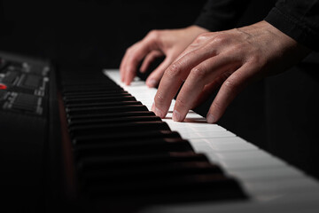 Fototapeta premium A pair of hands with long slender fingers gently presses down on the white keys of a digital piano illuminated by a soft dramatic light against a dark background.