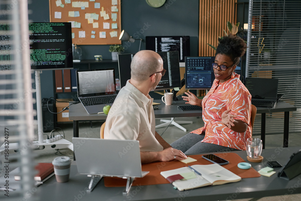 Poster black woman discussing project with caucasian middle aged man in modern office, multiple computer mo