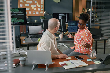 Black woman discussing project with Caucasian middle aged man in modern office, multiple computer...