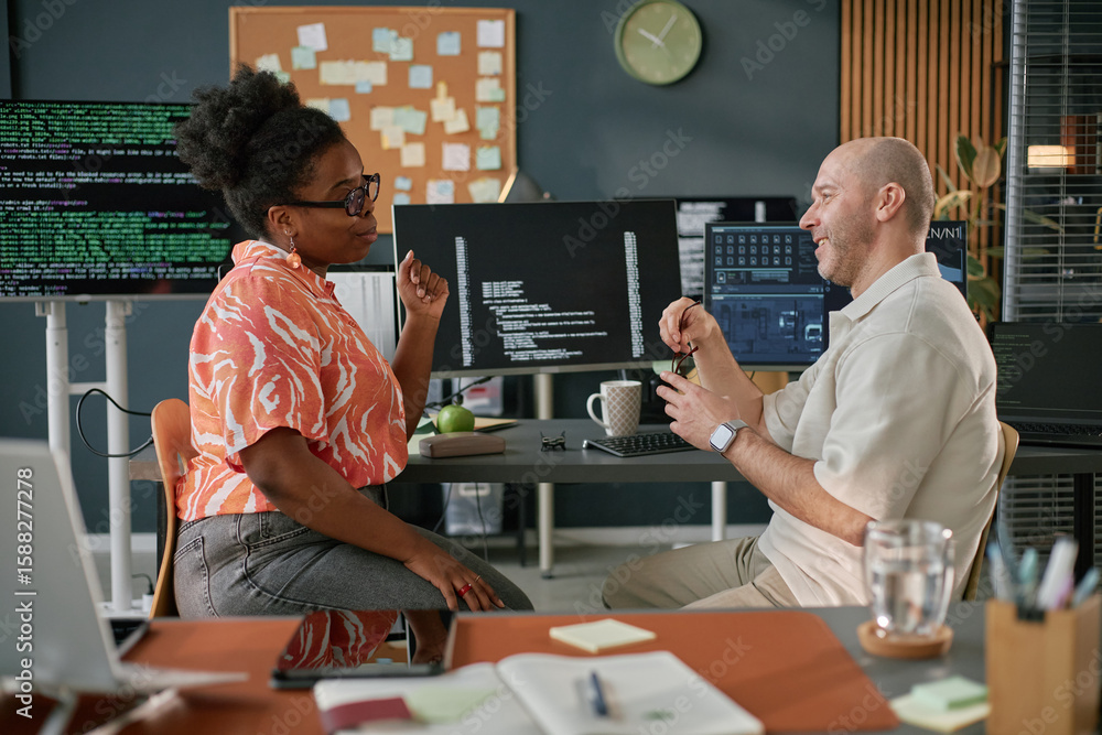 Poster black woman and caucasian middle aged man discussing programming code in modern office, both sitting