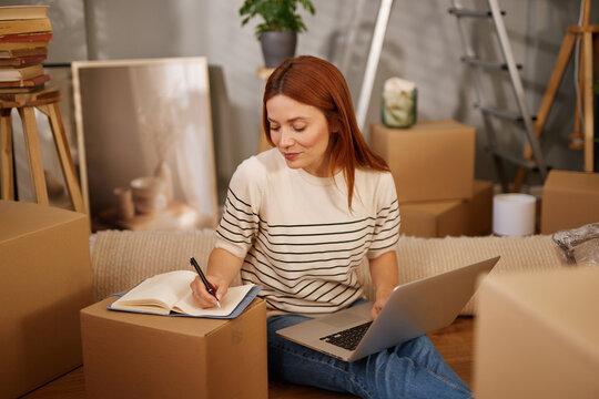 A woman sits on the floor surrounded by moving boxes. She writes notes in a notebook while using a laptop, focused on managing her move and ensuring everything is in order.
