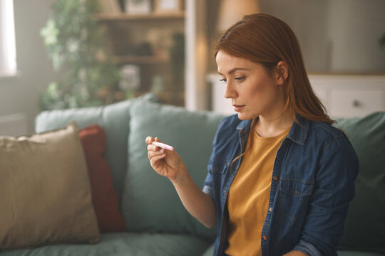 In a warm, inviting living room, a woman with red hair reviews the results of a pregnancy test, showcasing a mix of emotions as she contemplates her situation and future.