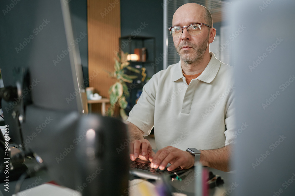 Poster portrait of middle aged caucasian man wearing glasses working at computer in modern office, typing o
