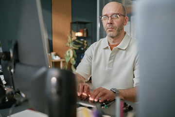 Portrait of middle aged Caucasian man wearing glasses working at computer in modern office, typing...