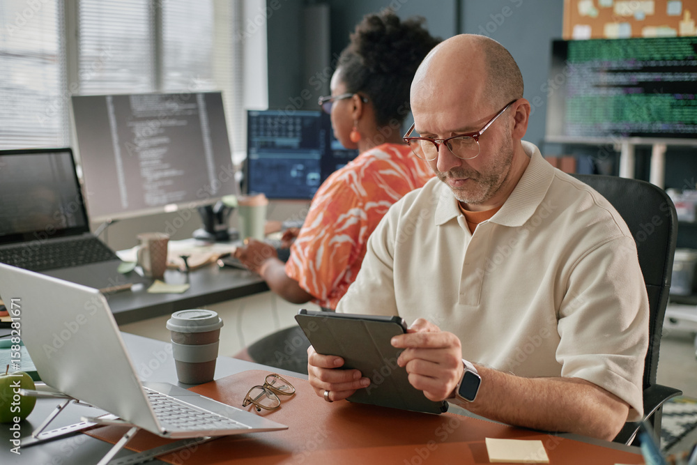 Poster middle aged caucasian man using digital tablet at desk while black woman working on computer in back