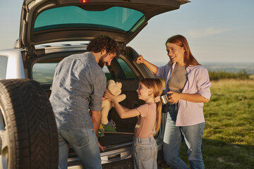 Family members prepare for a picnic by unloading a vehicle in a lush green countryside. They share...