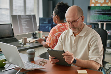 Middle aged Caucasian man using digital tablet at desk while Black woman working on computer in...