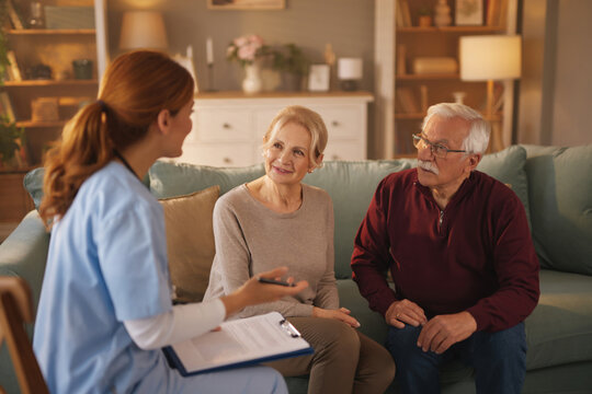 A home health aide, wearing scrubs and holding a clipboard, speaks to a senior couple sitting on a couch in their living room, discussing details of their care and needs. - Powered by Adobe