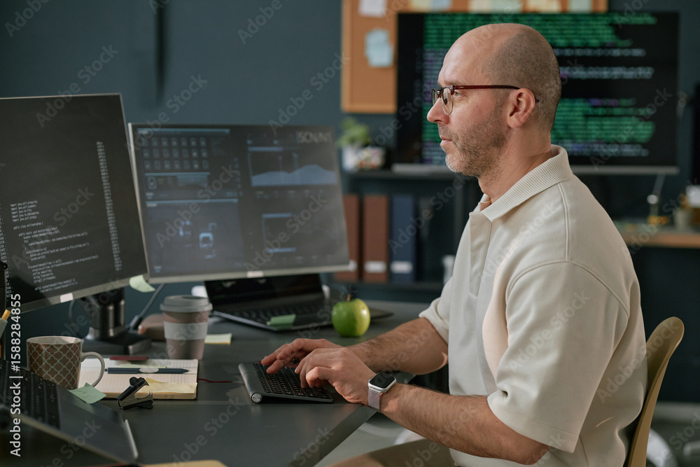 Poster middle aged caucasian man working at computer desk using keyboard and multiple monitors, wearing gla