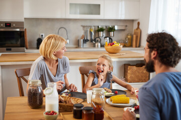 A family is gathered around a wooden table sharing breakfast in a sunlit kitchen. They are smiling...