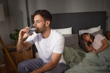 A man sits on the edge of a bed, sipping water from a glass while his partner rests peacefully in...