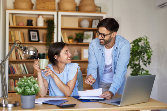 A man and a woman engage in a productive discussion at their home office workspace, enjoying coffee and reviewing documents on a laptop while surrounded by plants and books.