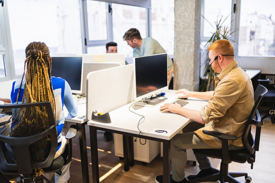 Diverse team of employees working on computers in a bright, open plan coworking space, collaborating and focusing on their tasks
