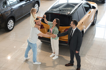 Salesman shaking hands with happy family near new car in salon