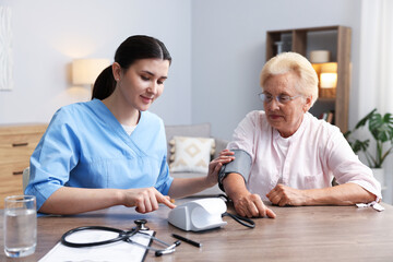 Nurse measuring patient's blood pressure at wooden table indoors. Home health care service