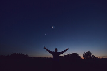 Silhouette of a man and countryside under the Moon, stars and planets in blue hour time.