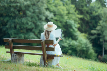 Elegant woman wearing summer clothes and hat reads peacefully on a bench in a nature landscape