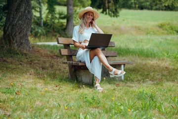 Calm nature backdrop as woman uses laptop on bench wearing summer outfit in rural environment
