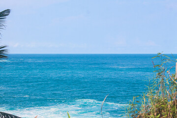 A tropical seaside view features the vibrant blue ocean and horizon framed by lush green foliage...