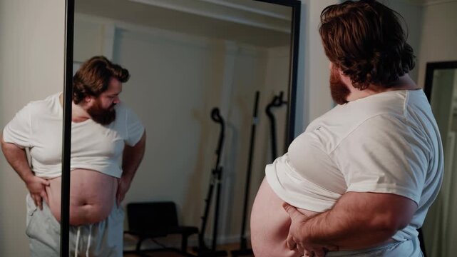 Fat man standing in front of mirror. Man is reflected in a mirror indoors. A woman in a casual shirt is examining her body for overweight self-observation. An overweight man positioned in lifestyle.