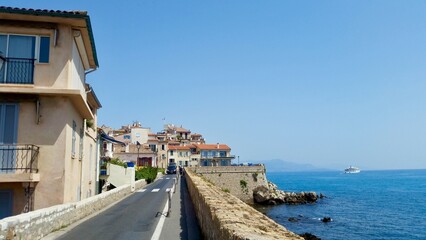 A charming coastal road in Antibes, France, with traditional buildings, while a historic stone wall borders the other, leading towards a distant town and a cruise ship.