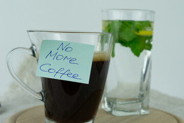 Black coffee cup with a sticky note saying No More Coffee next to a glass of water with mint leaves...